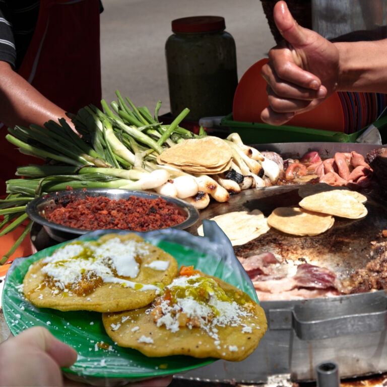 Close-up of fresh Mexican street food featuring garnachas with salsa and cheese on a green plate, with a vendor cooking on a traditional comal in the background.