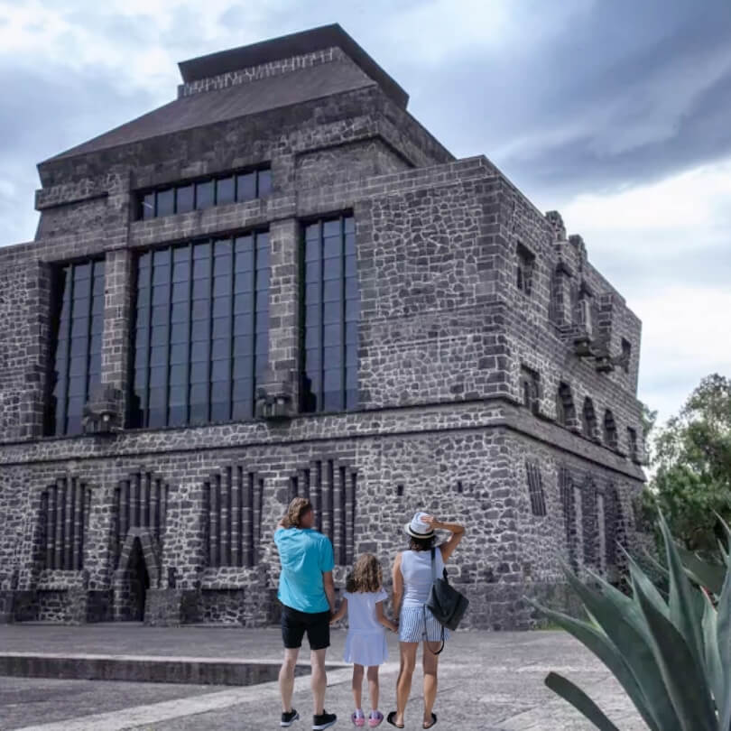 A family of three standing in front of the Anahuacalli Museum in Mexico City, admiring the volcanic stone architecture designed by Diego Rivera under a cloudy sky.