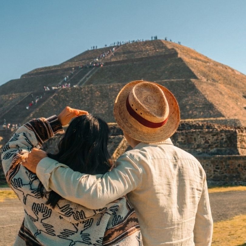 Back view of a couple in traditional travel attire looking at the ancient Pyramid of the Sun in Teotihuacán, Mexico, under a bright blue sky.