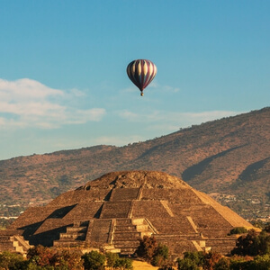 A colorful hot air balloon flying over the Pyramid of the Moon at the Teotihuacan archaeological site in Mexico during a clear morning.